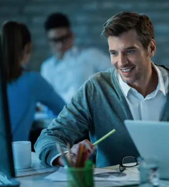 happy-male-entrepreneur-using-desktop-pc-while-working-office-there-are-people-background